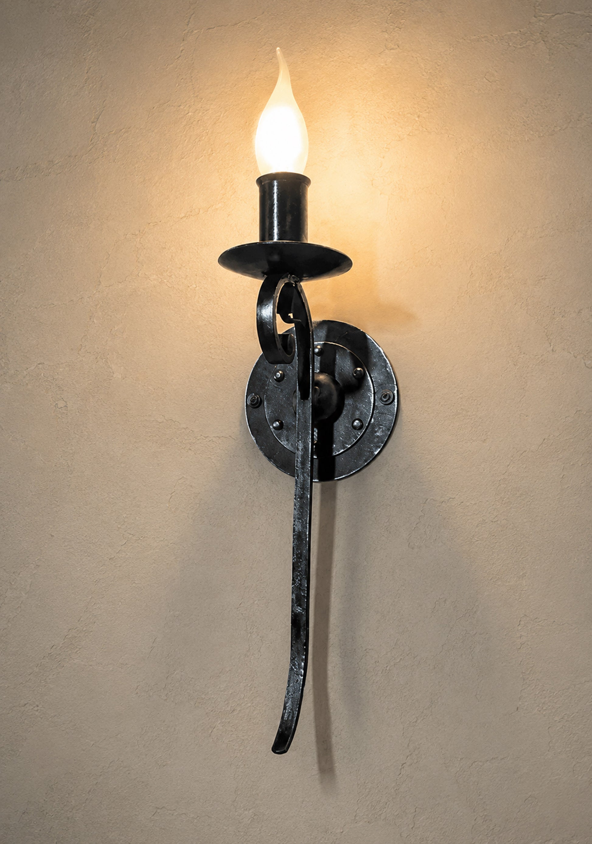 A black metal wall sconce with a curved gothic-style arm and a glowing flame-shaped bulb, mounted on a textured beige wall.