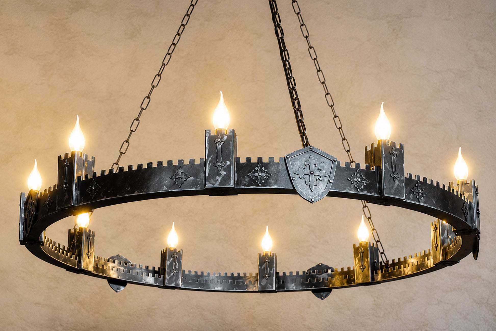 A medieval-style ring chandelier with a dark metal circular frame featuring castle-tower motifs, a central shield emblem, and multiple candle-style lights glowing against a textured beige wall.