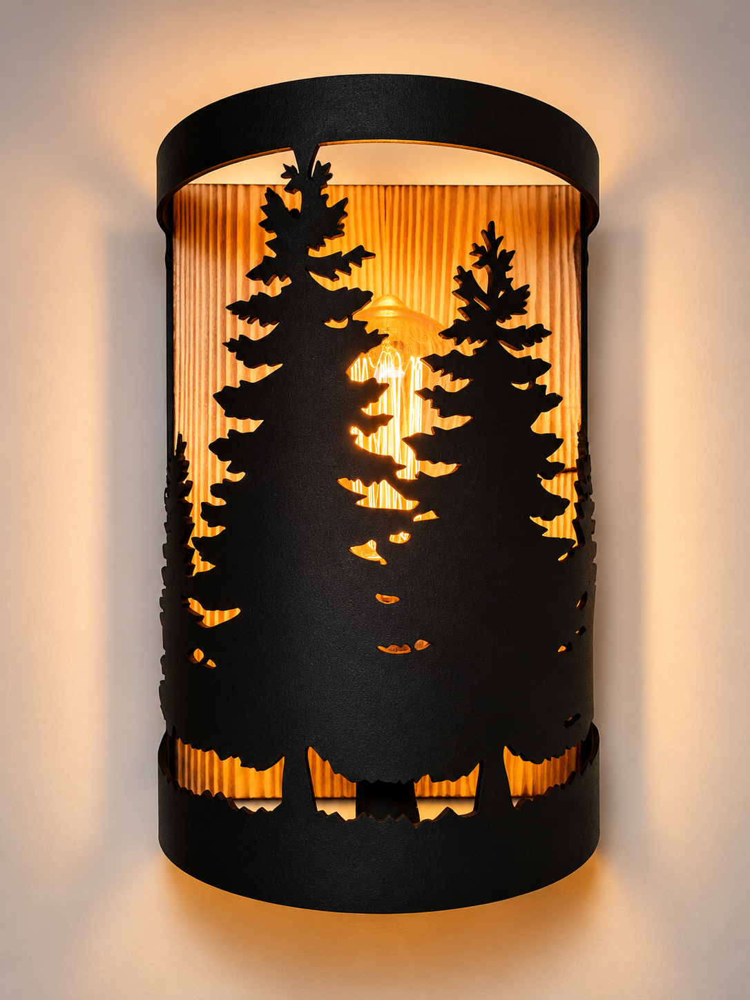 A cylindrical wall sconce with a black metal frame featuring laser-cut silhouettes of pine trees, illuminated from within by a warm yellow light that casts a sunset-like glow on a beige wall.
