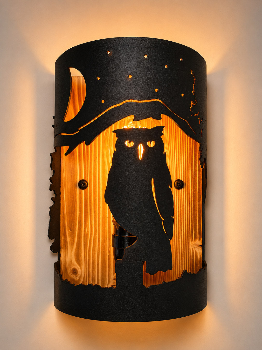 A cylindrical wall sconce light fixture featuring a black metal cutout design of an owl under a crescent moon and stars, set against a warm glowing wood-grain background.