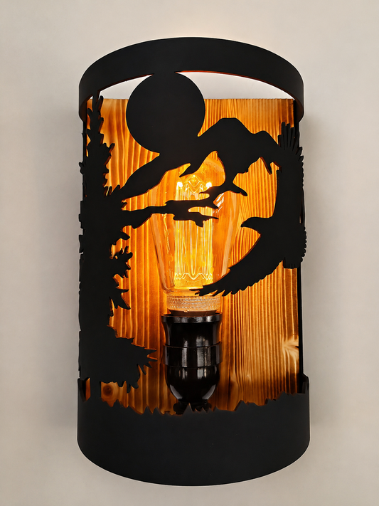 A wall sconce with a black metal silhouette design of a tree, moon, and flying birds over a warm orange-brown wood-grain background, illuminated by an exposed filament light bulb.